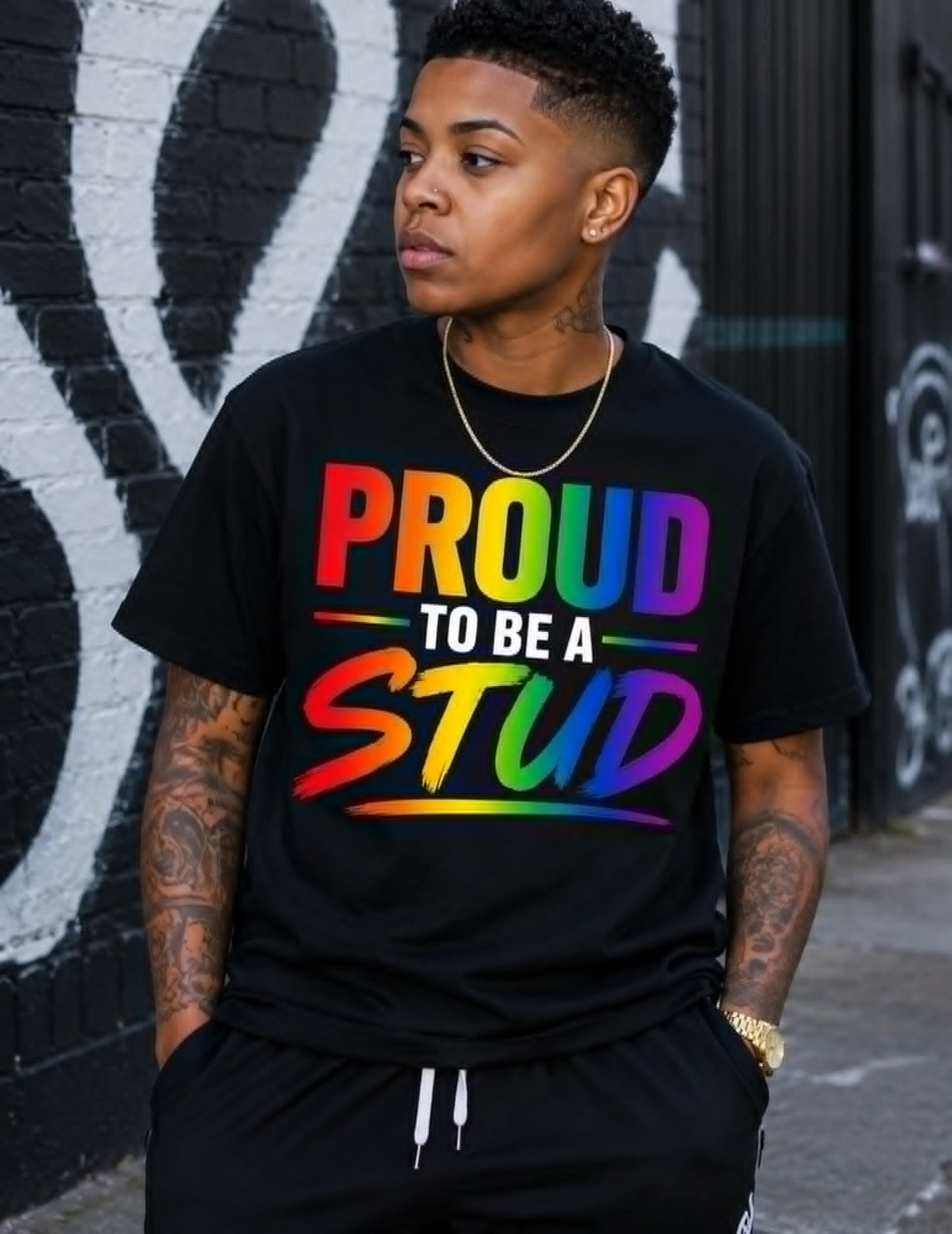 Person wearing a black t-shirt with colorful text 'PROUD TO BE A STUD' against a graffiti-covered wall.